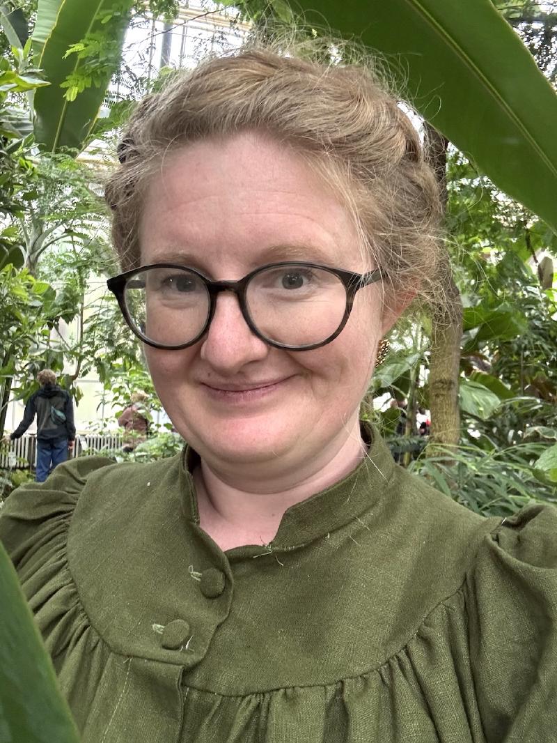 Tight shot of blonde white woman age 42, dark round plastic glasses, braided hair, in a green high-collared dress, smiling slightly and looking directly at the camera with palm trees in a conservatory in the background
